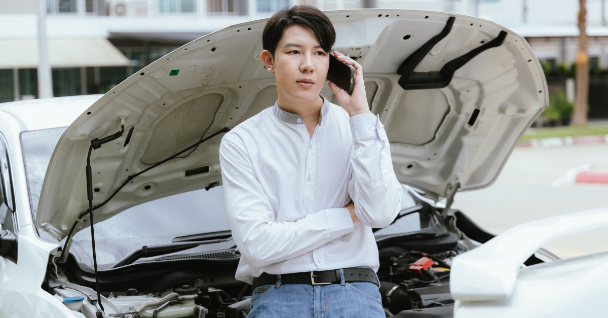 A bearded man in his 30s stands beside his broken-down car